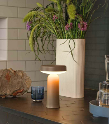Lampă de masă portabilă Ease de la Muuto pe un blat de bucătărie închis la culoare, decorat cu flori.