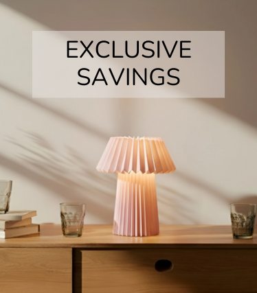 Lampă de masă roz pal pe o măsuță din lemn, înconjurată de pahare și cărți în lumina caldă a soarelui.