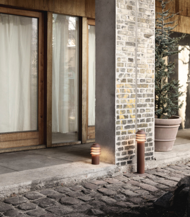 Scenă outdoor cu două lămpi moderne, cilindrice, de iluminat alei, amplasate lângă o potecă din piatră și un stâlp din cărămidă și beton. În spate se văd uși din sticlă cu ramă din lemn, perdele ușoare și o plantă evergreen mare în ghiveci. Lumina caldă a lămpilor creează o atmosferă confortabilă și primitoare.