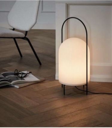 Lampă albă striată cu cadru negru tip mâner pe parchet în model herringbone, lângă un fotoliu.